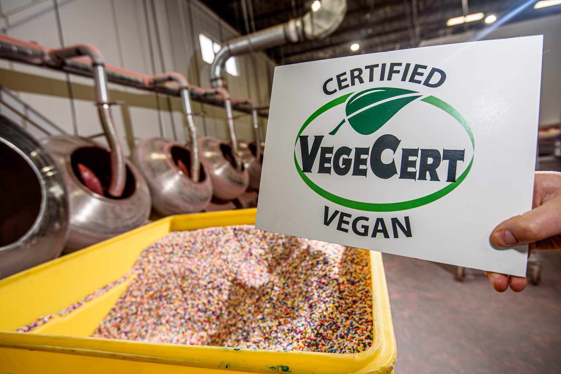 A hand holding a Certified VegeCert Vegan seal inside a candy production facility